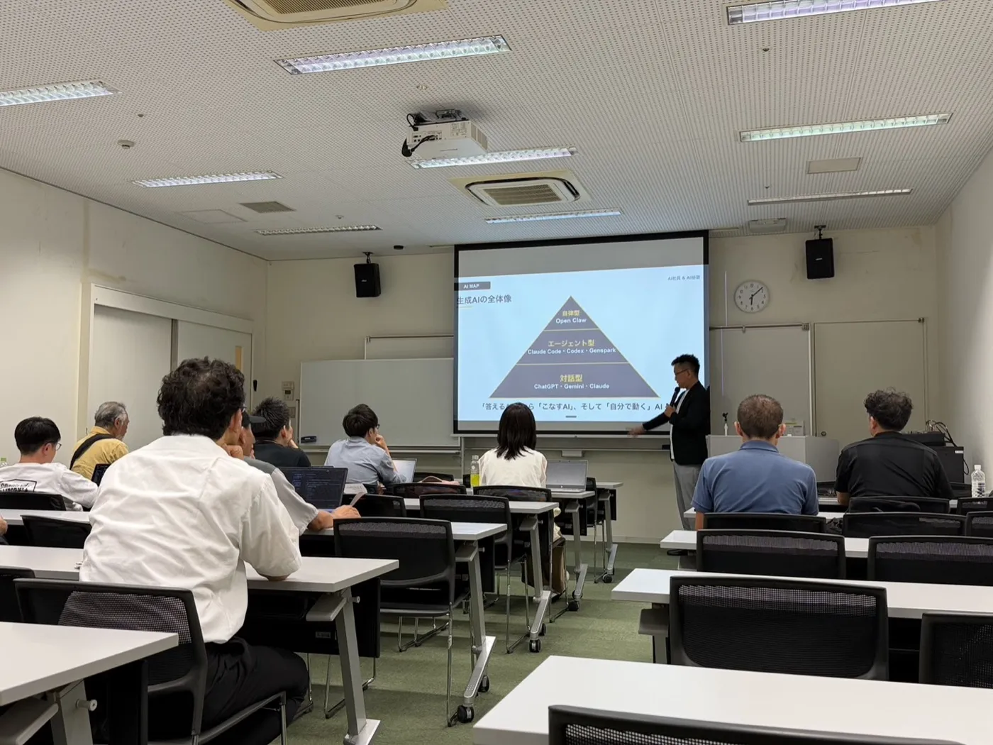 会場全体を後方から撮影した引きの写真。参加者が着席し、前方のスクリーンに講師のスライドが投影されています